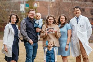 Group photo of Heidi Neas holding an ultrasound photo, standing with her husband, two children and three medical professionals