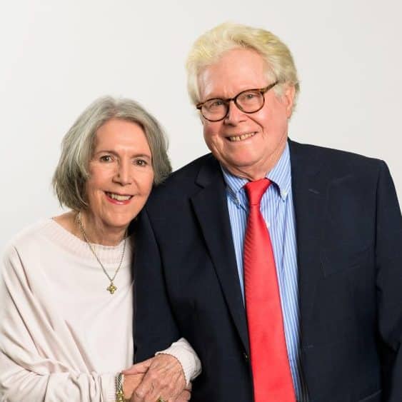 An elderly man in glasses and a black suit with red tie - Tom Michaels – stands beside his wife, Judy Michaels.