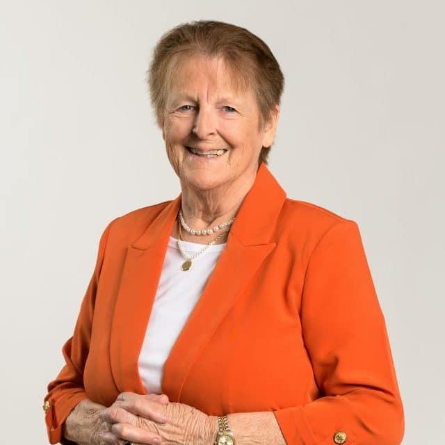 An elderly woman – Debby Davis – wearing a white shirt and an orange dress jacket.