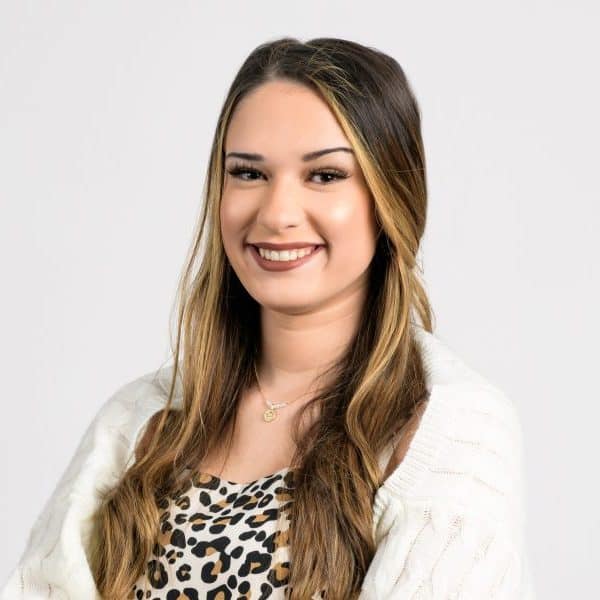 A young woman – Selena Avalos – in a white coat and a leopard print dress, smiling to camera.