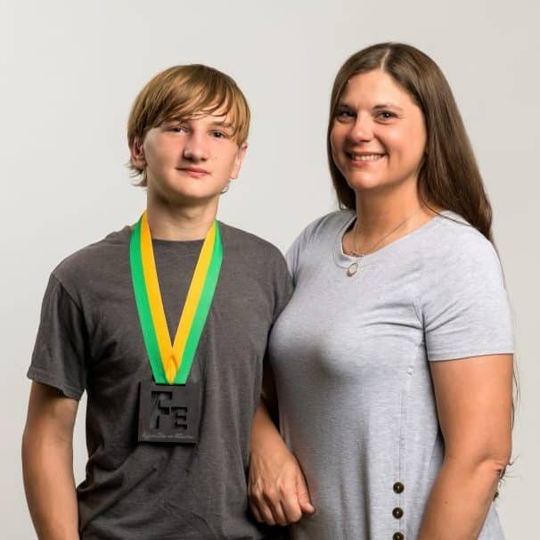 A teenage boy in a grey t-shirt – Hunter Creamer – wearing the "Fe" Iron Strong medal; standing next to him is his mother, Marilyn Casas.
