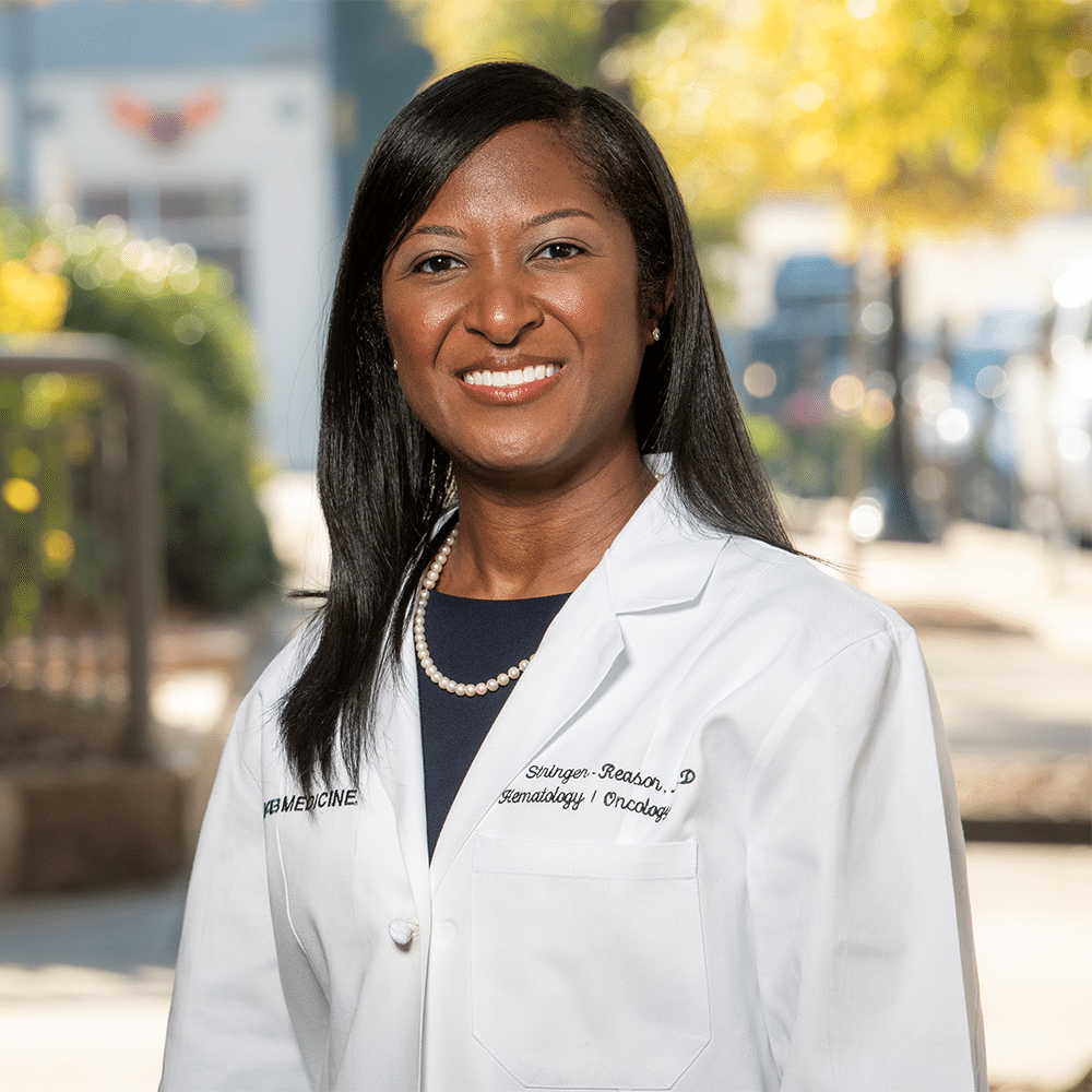 A headshot of Erica Stringer-Reasor, M.D.; A Black woman in a lab coat stands outdoors, smiling to camera.