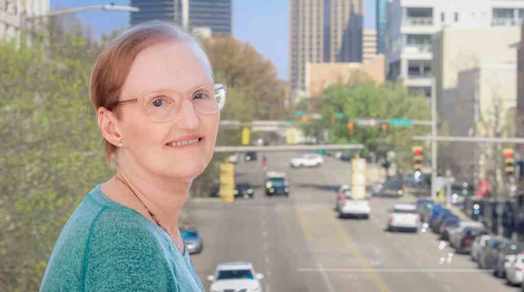 A headshot of cancer patient Rebecca Hall. Rebecca, a middle-aged woman, stands smiling to camera standing above a busy street in Birmingham, AL.