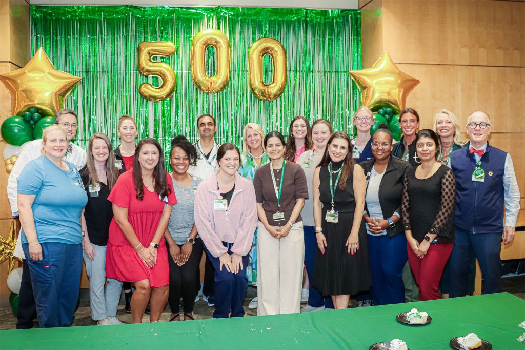 Another photo of the UAB O'Neal Cancer Center Patient Service Line celebrating the 500th Car T patient, now joined by Barry Sleckman, M.D., Ph.D.; the group stands in a wood-walled room behind a green table in front of balloons reading "500."
