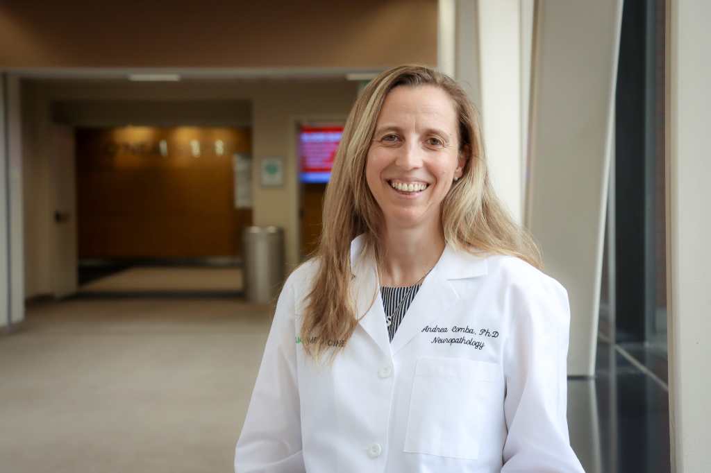 A woman in a white lab coat stands in a hallway outside the O'Neal Cancer Center at UAB Hospital – this is Andrea Comba, Ph.D.