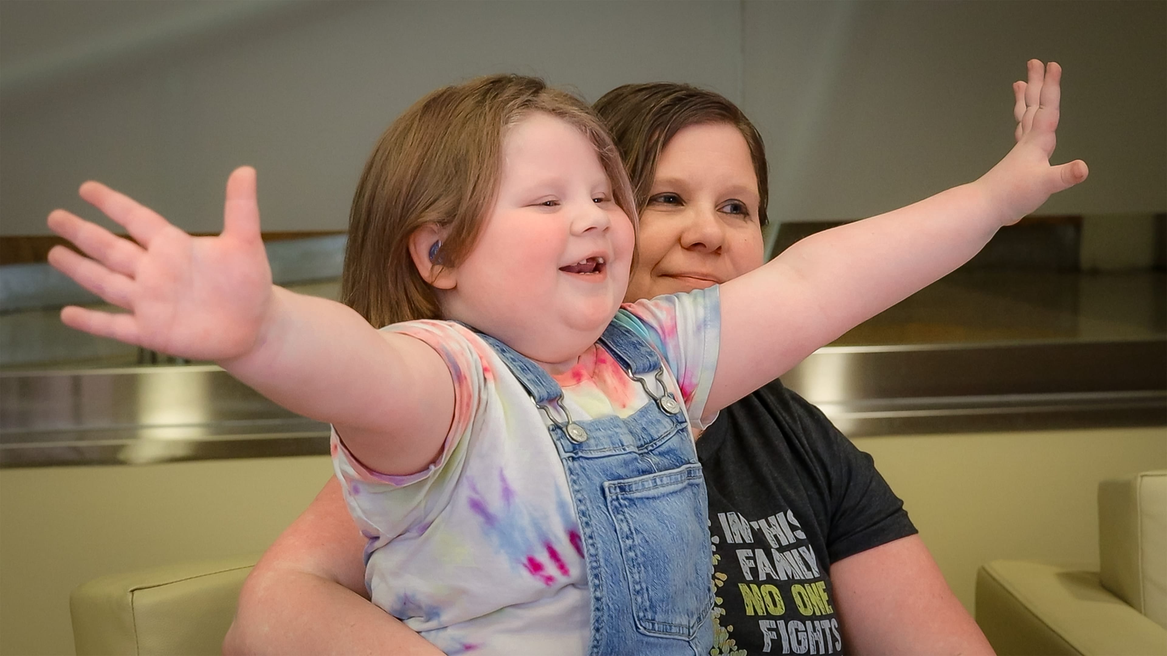 Piper Diamond, a cancer survivor and Iron Strong honoree, smiles to camera and holds her arms our wide; her mother, Rhonda, sits behind her, smiling.