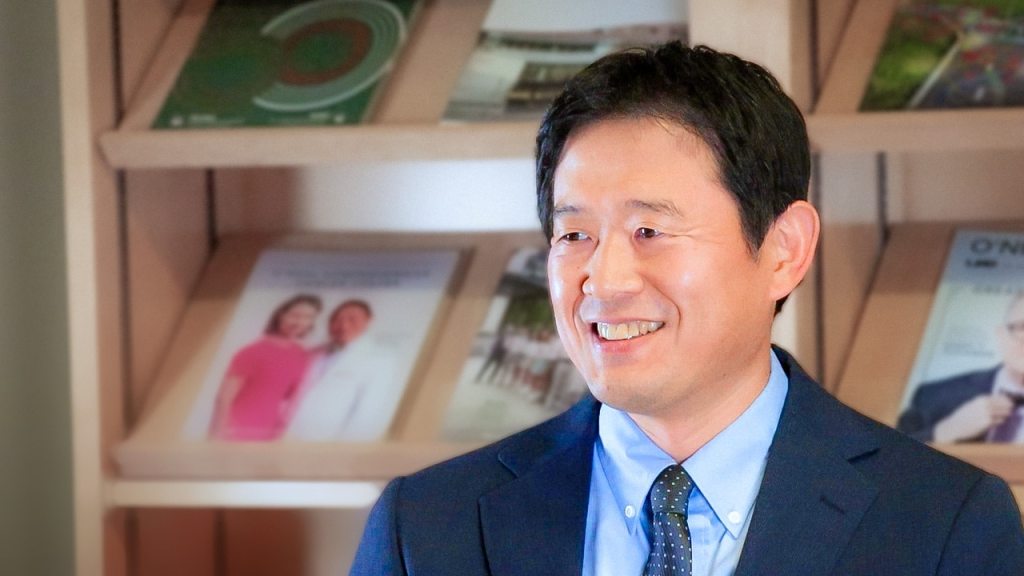 A headshot of Satoru Osuka, M.D., Ph.D., against a pair of bookshelves.