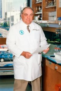 A man in a white lab coat with a UAB Comprehensive Cancer Center logo stands in a laboratory, with the city of Birmimgham visible in a window behind him; Albert F. LoBuglio, M.D.