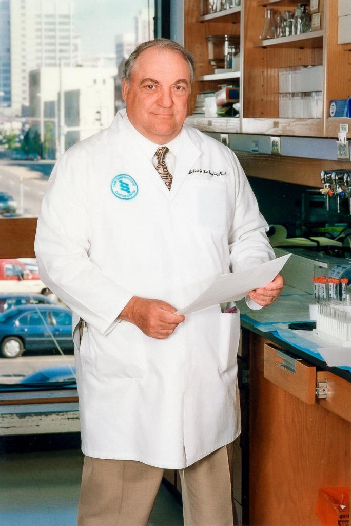 A man in a white lab coat with a UAB Comprehensive Cancer Center logo stands in a laboratory, with the city of Birmimgham visible in a window behind him; Albert F. LoBuglio, M.D.